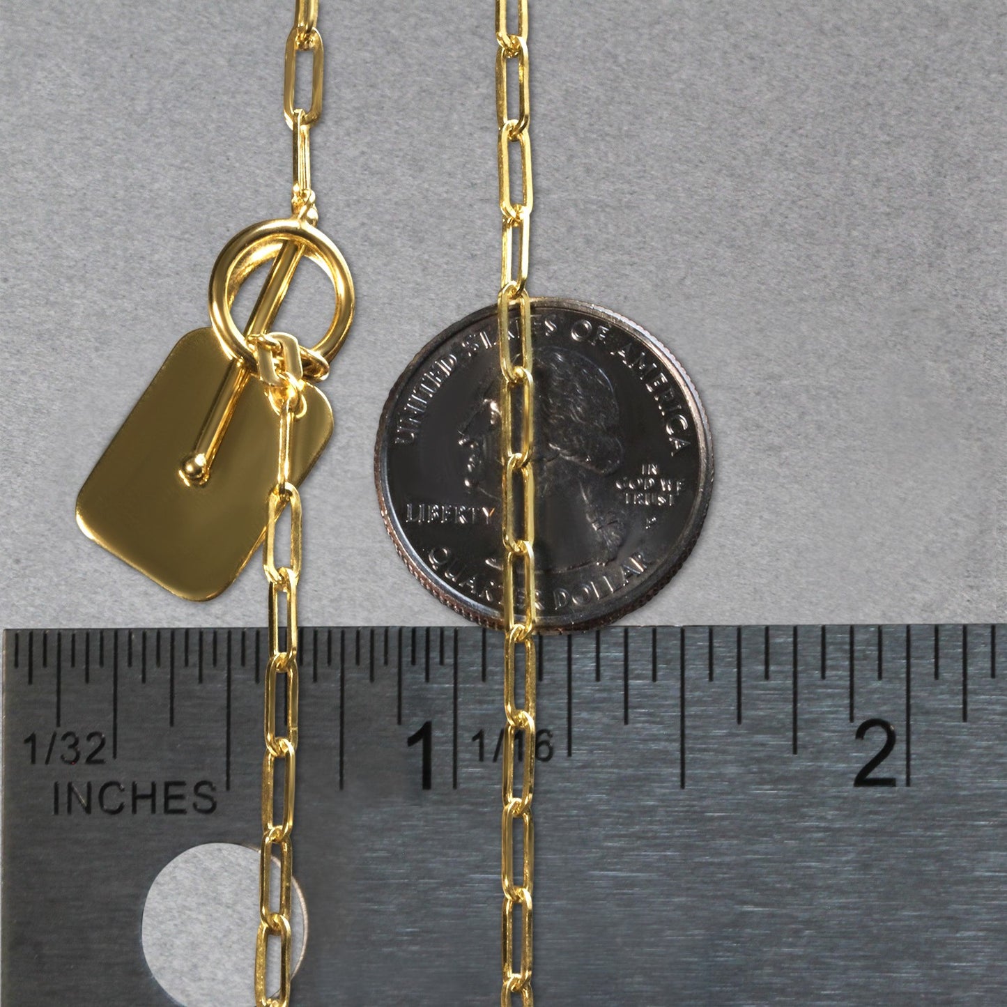 Gold chain with a tag and clasp next to a ruler for size reference on a gray background