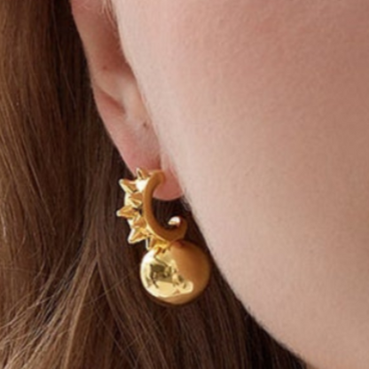 Close-up of a gold sunburst hoop earring with a spherical drop, worn on a woman’s ear